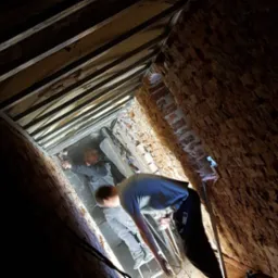 Dwóch mężczyzn pracuje w wąskiej przestrzeni między ceglanymi ścianami i drewnianą konstrukcją sufitu, jeden z młotkiem mocuje element konstrukcji, drugi stoi na drabinie, oświetlenie punktowe.