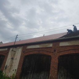 REMS & STEL DYNAMICS - Dwóch pracowników na dachu starego budynku z czerwonej cegły, układających czerwoną dachówkę na tle pochmurnego nieba.