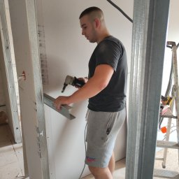 REMS & STEL DYNAMICS - Pracownik w szarych spodenkach i czarnej koszulce montuje metalowy profil do płyty gipsowo-kartonowej za pomocą elektrycznego nożyka do blachy, widoczny fragment drabiny i instalacja elektryczna.