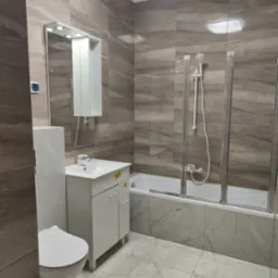 Nowoczesna łazienka z płytkami imitującymi marmur na podłodze i ścianach, biała szafka pod umywalką z lustrem z oświetleniem LED, toaleta i wanna z parawanem nawannowym.