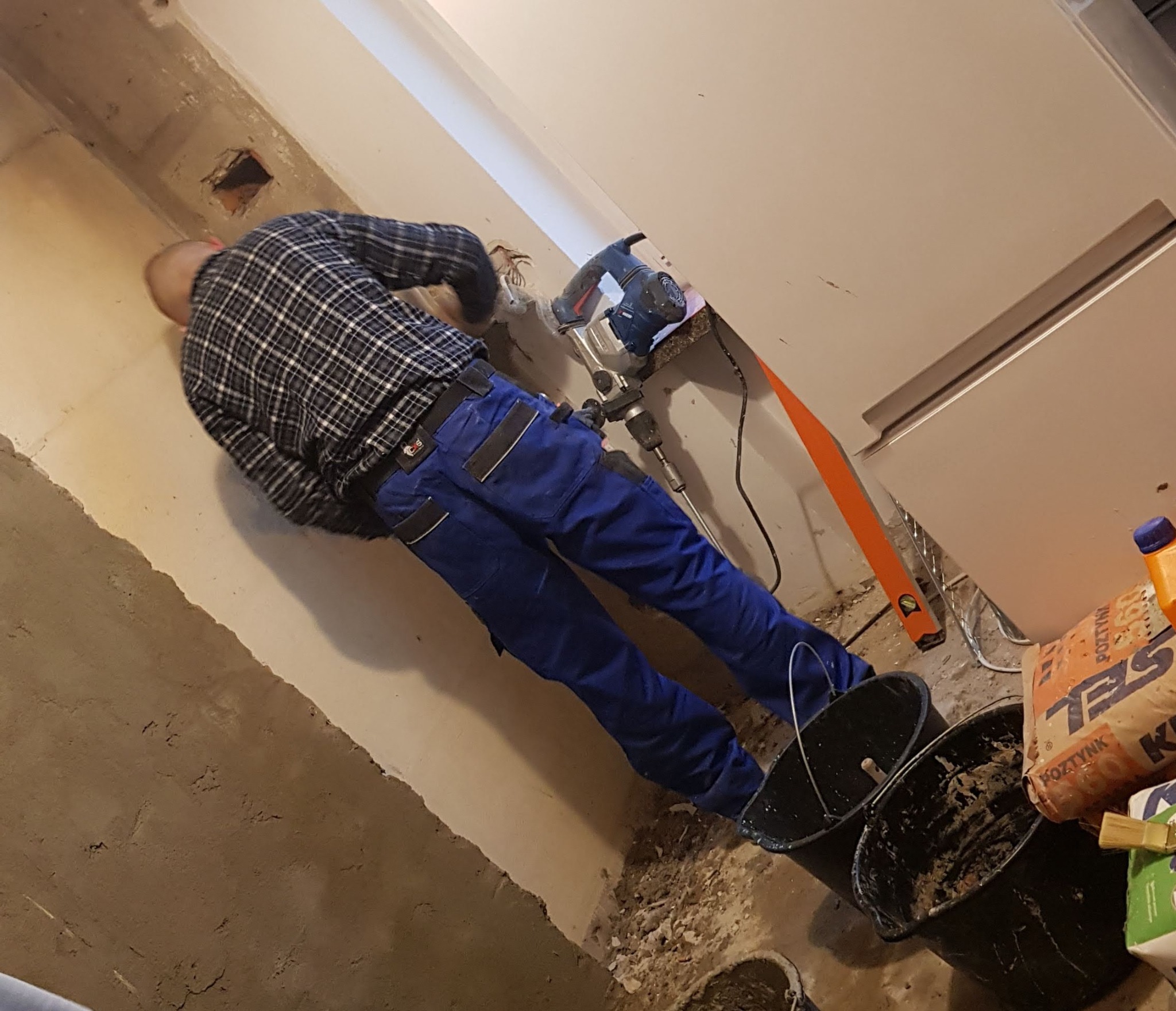 Pracownik w niebieskich spodniach i kraciastej koszuli używa młota udarowego do kucia ściany podczas remontu, widoczne wiadra, poziomica i worek z zaprawą.
