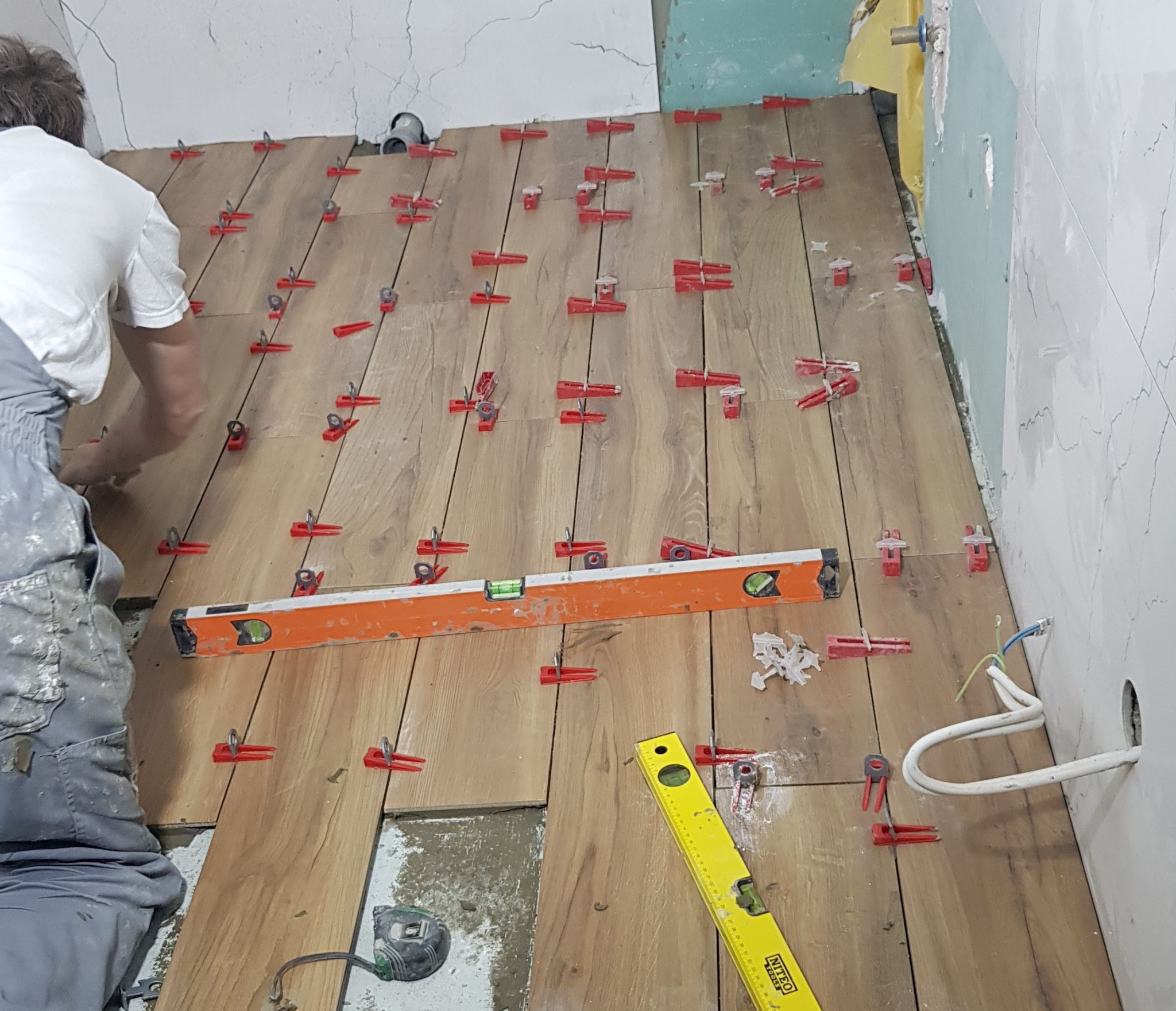 Układanie paneli podłogowych imitujących drewno z użyciem czerwonych klipsów poziomujących; widoczne poziomice, częściowo wykończona ściana i wystające kable elektryczne.
