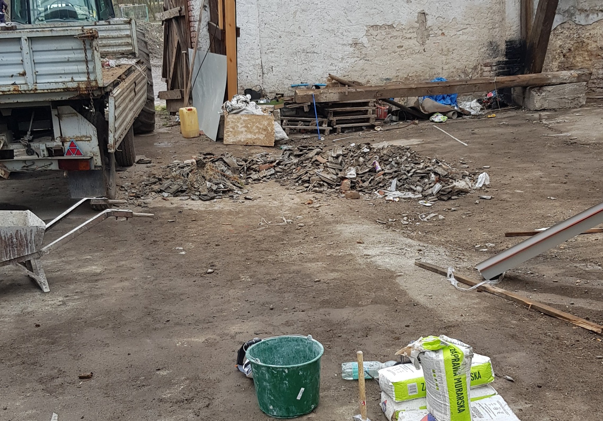 Sterta gruzu i resztek materiałów budowlanych na placu budowy, obok zielone wiadro, worki z zaprawą murarską i taczka, w tle zaparkowany stary traktor.