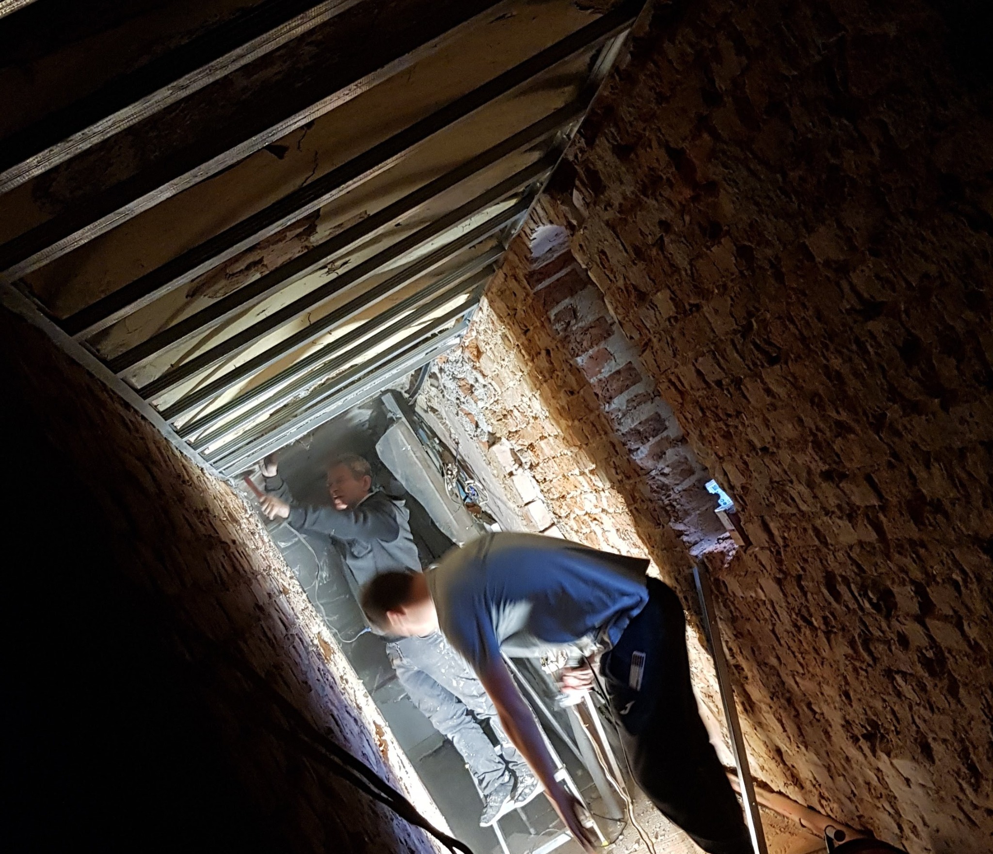 Dwóch mężczyzn pracuje w wąskiej przestrzeni między ceglanymi ścianami i drewnianą konstrukcją sufitu, jeden z młotkiem mocuje element konstrukcji, drugi stoi na drabinie, oświetlenie punktowe.