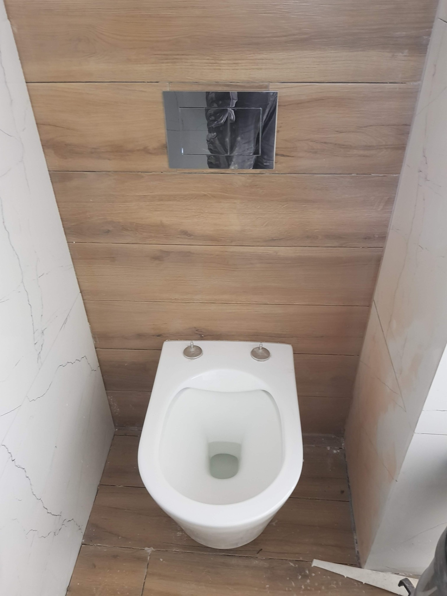 Podwieszana toaleta z chromowanym przyciskiem spłukującym na ścianie wyłożonej panelami drewnopodobnymi, obok ściana z marmurowymi płytkami, widoczne niedociągnięcia w wykończeniu.
