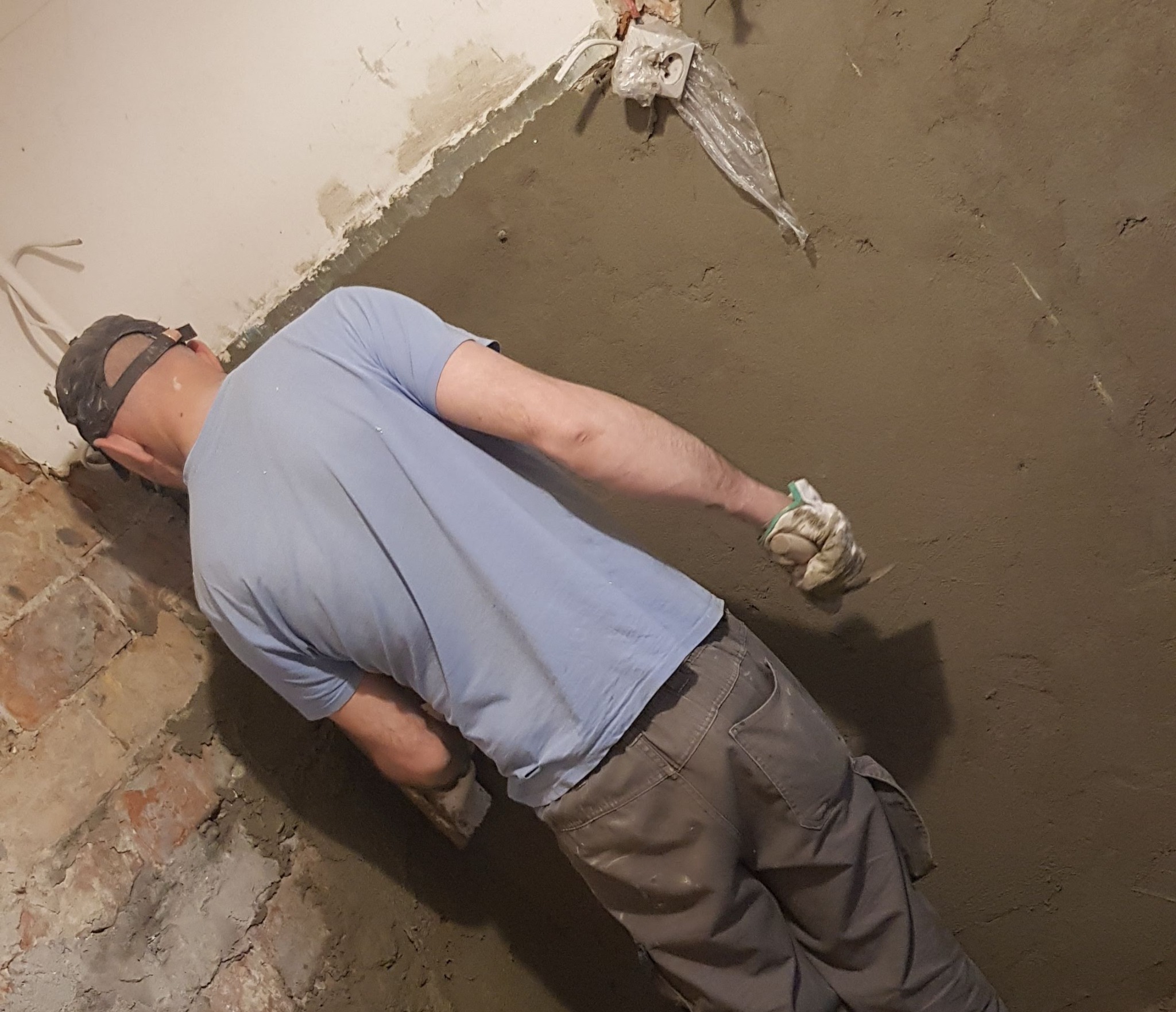 Mężczyzna w czapce i roboczych spodniach nakłada szarą masę szpachlową na ścianę z częściowo odsłoniętą czerwoną cegłą, zabezpieczone gniazdko elektryczne w tle.