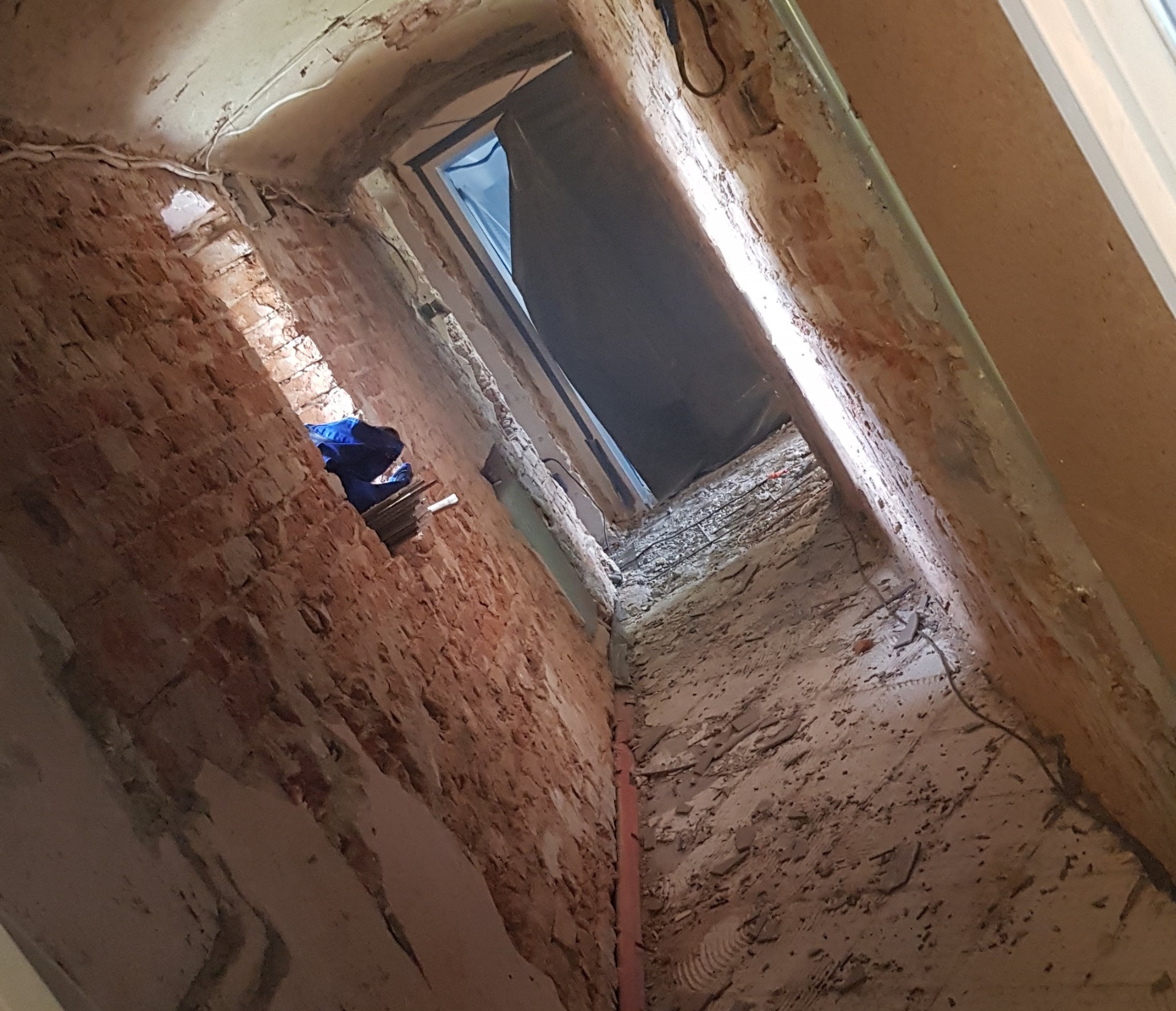 Wnętrze w trakcie remontu: surowe cegły, fragment nieotynkowanej ściany, widoczne przewody elektryczne i rura pionowa, gruz na podłodze, otwór drzwiowy z zasłoną.