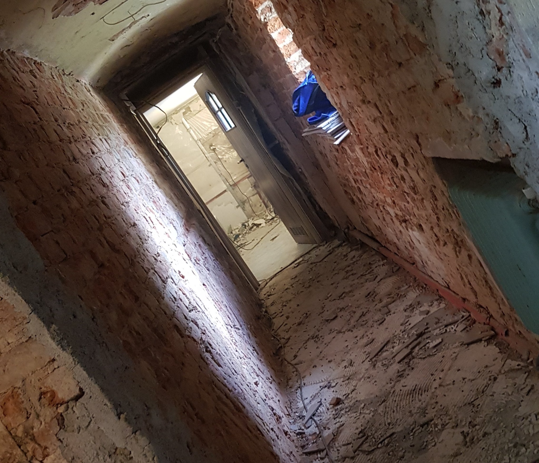 Wnętrze w trakcie remontu z odsłoniętą cegłą, widoczne ślady po skuciu tynku, drzwi w głębi z widokiem na pomieszczenie z gruzem.