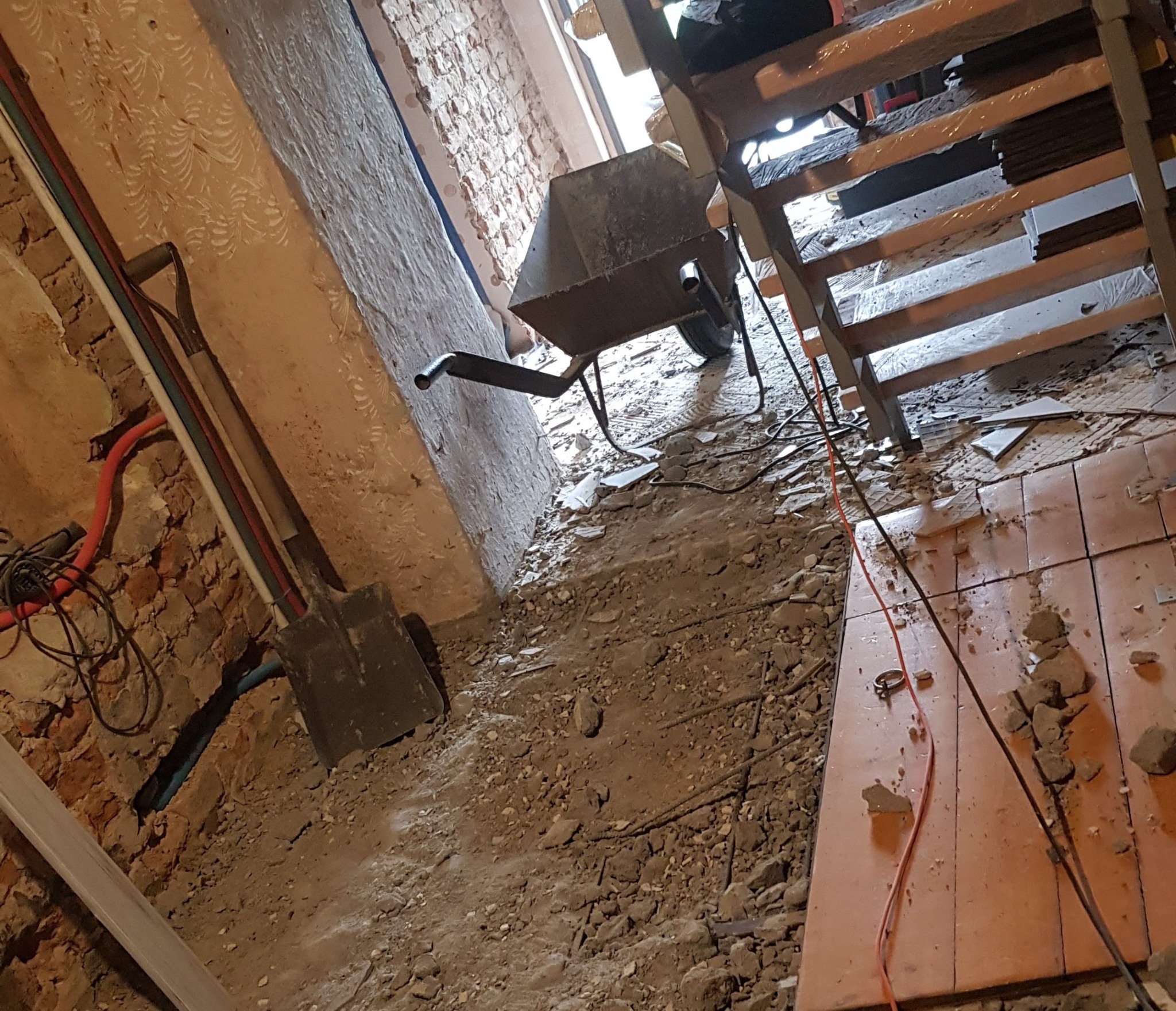 Wnętrze w trakcie demontażu podłogi, widoczna stara ceglana ściana, gruz, łopata oparta o ścianę, taczka i drewniane schody.