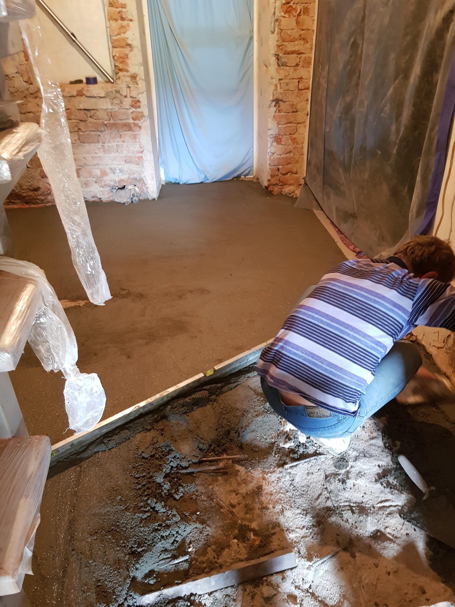 Mężczyzna w koszuli w paski wyrównuje wylewkę podłogową za pomocą poziomicy w pomieszczeniu z odsłoniętą cegłą i folią ochronną.