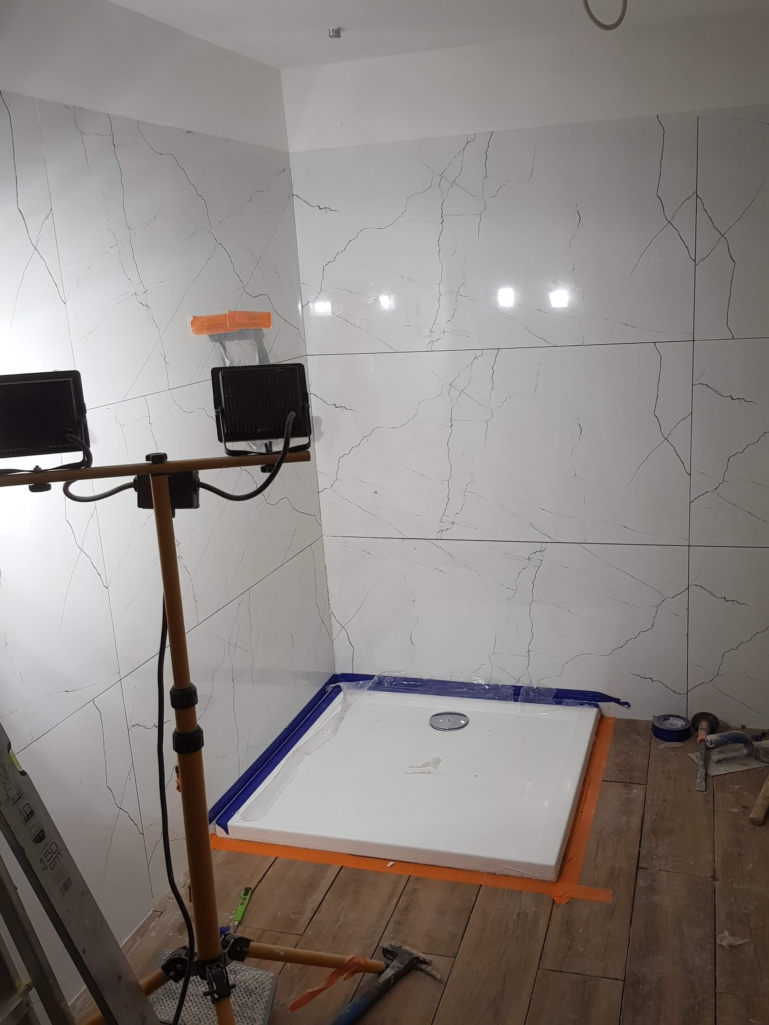 Biały brodzik zabezpieczony taśmą, otoczony płytkami imitującymi marmur, oświetlony lampami budowlanymi podczas prac wykończeniowych w łazience.