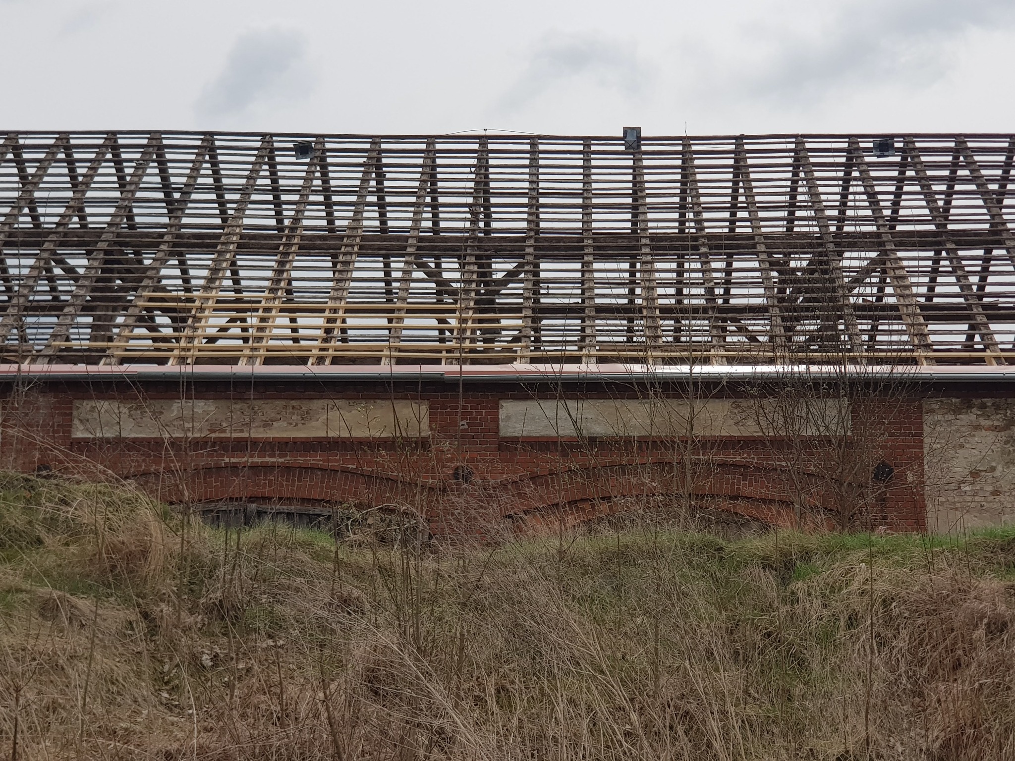 Konstrukcja dachu starego, ceglanego budynku w trakcie remontu, widoczna spod poziomu gruntu, z elementami drewnianej więźby i częściowo zdjętym pokryciem.
