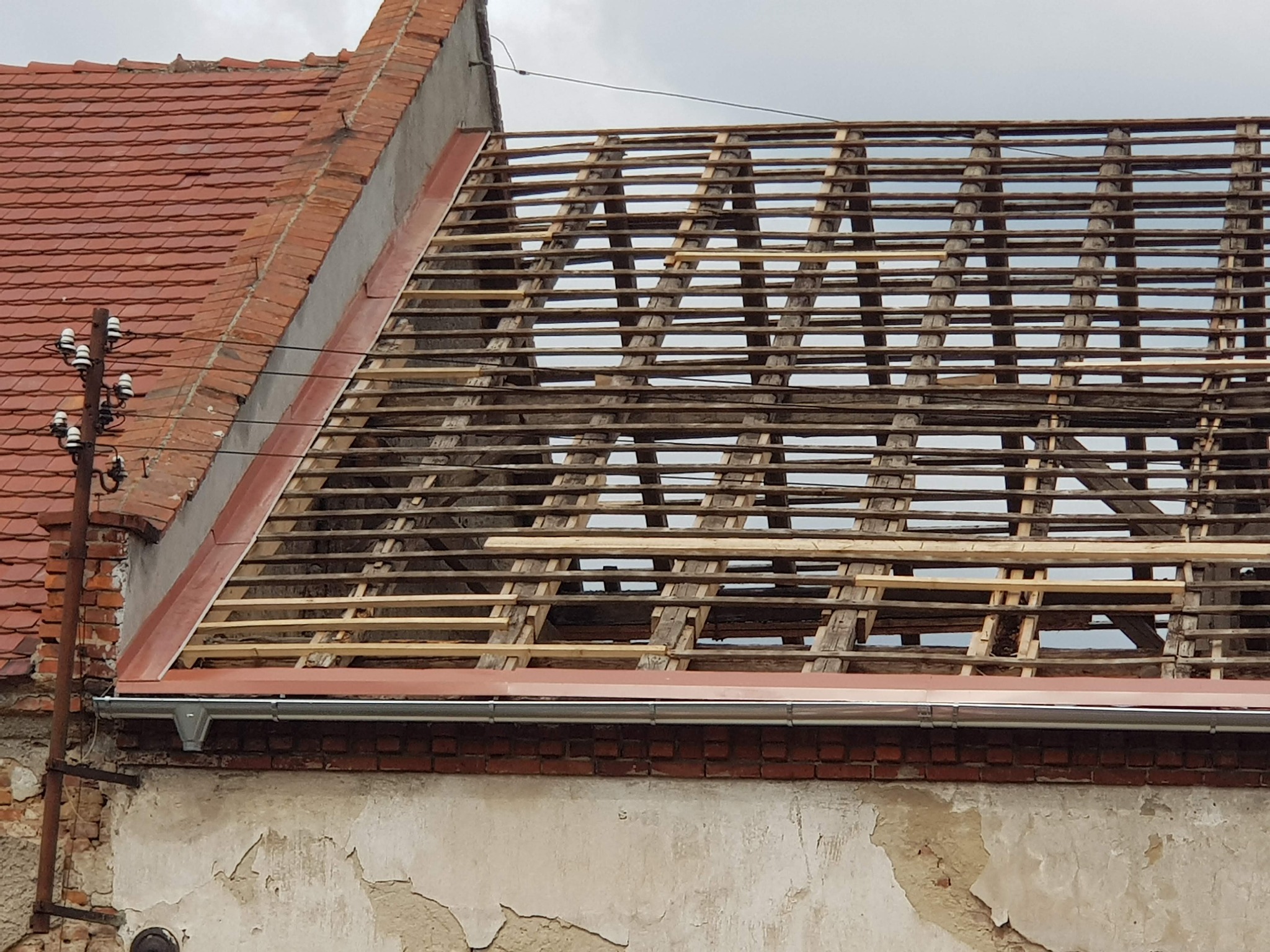 Konstrukcja dachu z widocznymi belkami, częściowo pokryta czerwoną dachówką, z widocznym fragmentem elewacji z odpadającym tynkiem i rynną.