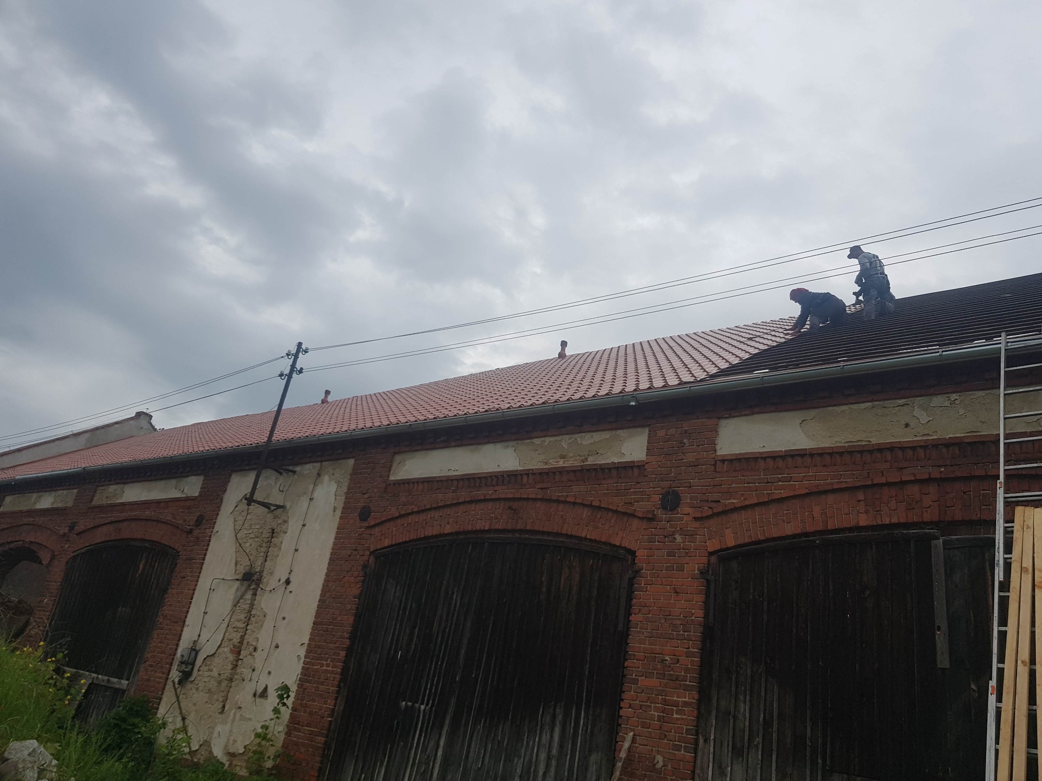 Dwóch pracowników na dachu starego budynku z czerwonej cegły, układających czerwoną dachówkę na tle pochmurnego nieba.