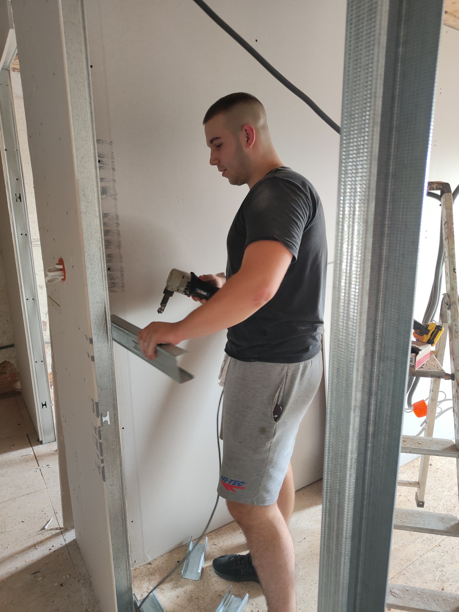 Pracownik w szarych spodenkach i czarnej koszulce montuje metalowy profil do płyty gipsowo-kartonowej za pomocą elektrycznego nożyka do blachy, widoczny fragment drabiny i instalacja elektryczna.