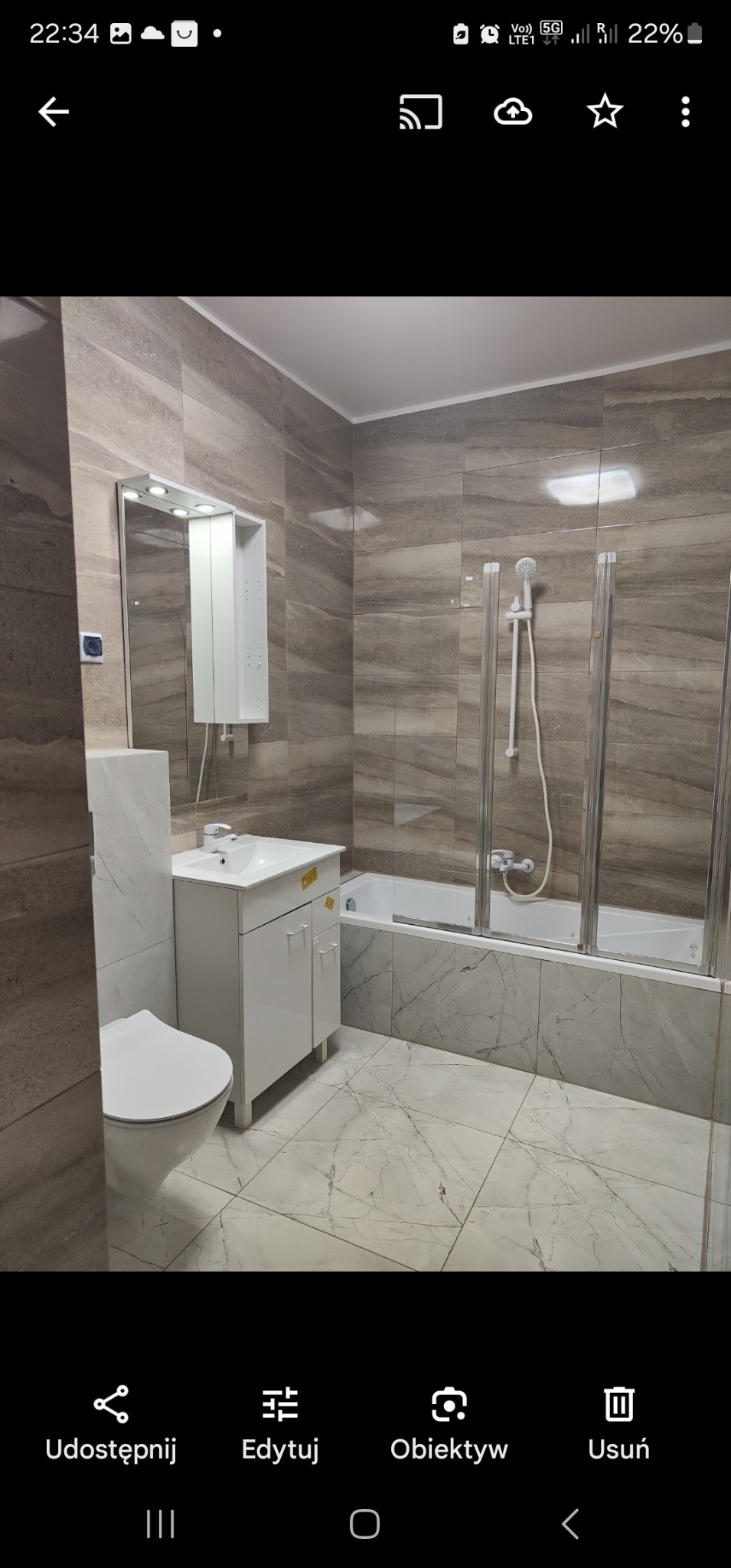 Nowoczesna łazienka z płytkami imitującymi marmur na podłodze i ścianach, biała szafka pod umywalką z lustrem z oświetleniem LED, toaleta i wanna z parawanem nawannowym.