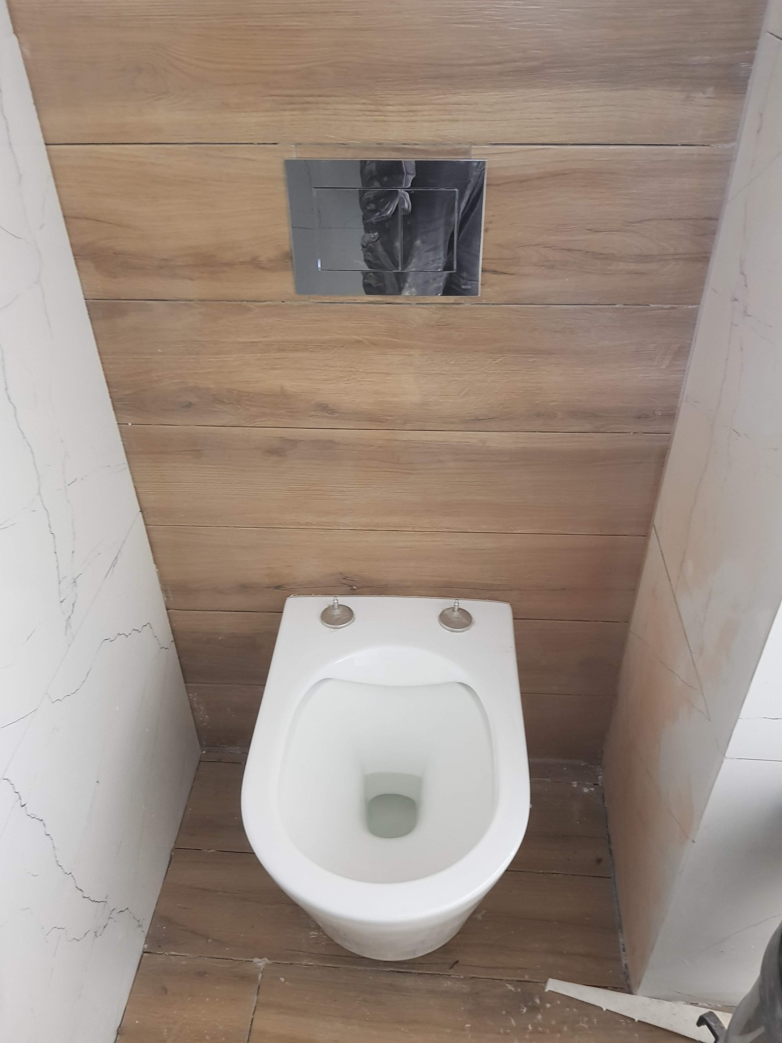 Wisząca toaleta z chromowanym przyciskiem spłukującym na ścianie wyłożonej panelami imitującymi drewno, obok ściany z imitacją marmuru, widoczne niedociągnięcia w wykończeniu podłogi.