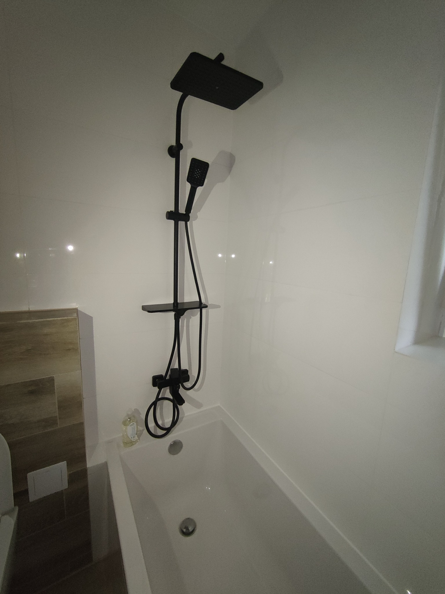 Nowoczesna łazienka z białą wanną i czarną armaturą prysznicową z deszczownicą, widok z góry na wannę i fragment ściany z błyszczącymi białymi płytkami.