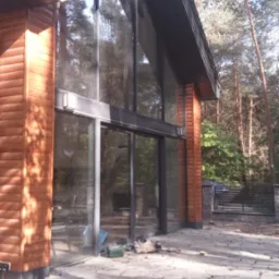 Nowoczesny dom z drewnianą elewacją i dużymi przeszkleniami, widoczne narzędzia i materiały budowlane na zewnątrz, sugerujące prace wykończeniowe przy tarasie lub elewacji.