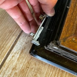 XBOOM SPÓŁKA Z OGRANICZONĄ ODPOWIEDZIALNOŚCIĄ - Detail ujęcia ręki przytrzymującej metalowy element regulacyjny w brudnej, drewnianej ramie okiennej w Bielsko-Białej, z widocznymi śladami kurzu i zabrudzeń.