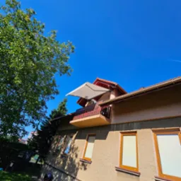 Zewnętrzny widok domu z beżową elewacją, drewnianymi oknami i balkonem z rozłożoną markizą w paski, sfotografowany w słoneczny dzień z perspektywy żabiej.