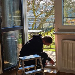 XBOOM SPÓŁKA Z OGRANICZONĄ ODPOWIEDZIALNOŚCIĄ - Mężczyzna w czarnej bluzie, klęczący na drabince, zajmuje się uszczelnianiem okna balkonowego z widokiem na ogród i metalową barierkę balkonu.