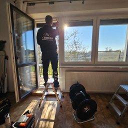 XBOOM SPÓŁKA Z OGRANICZONĄ ODPOWIEDZIALNOŚCIĄ - Mężczyzna w uniformie firmy XBOOM stojący na aluminiowej drabinie podczas regulacji okna PCV, widok z wnętrza mieszkania na drzewa i błękitne niebo.