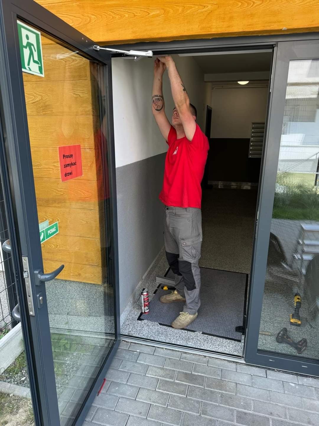 Pracownik w czerwonej koszulce reguluje samozamykacz w szarych drzwiach wejściowych do budynku, widoczna naklejka z instrukcją zamykania, narzędzia i piana montażowa obok progu.