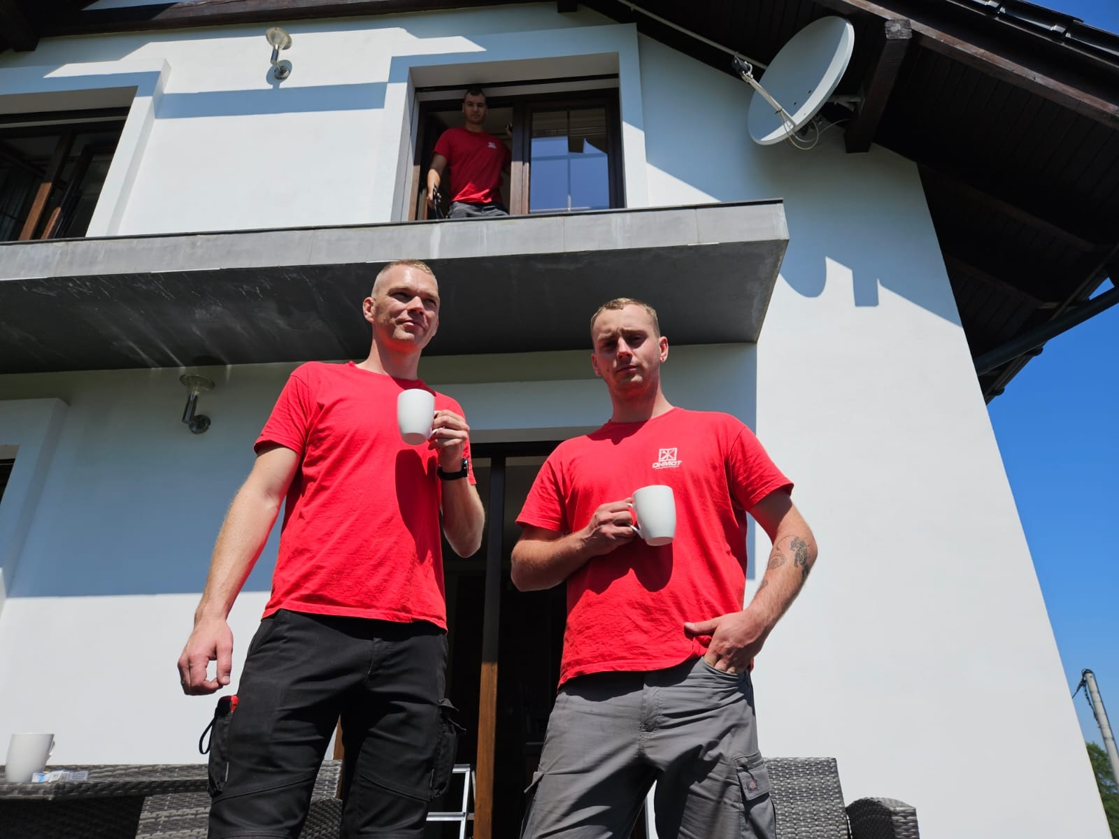 Trzech mężczyzn w czerwonych koszulkach, z kubkami w dłoniach, stoi przed domem z białymi ścianami i ciemnymi oknami, jeden z nich jest w oknie na piętrze. Na dachu widoczna antena satelitarna.