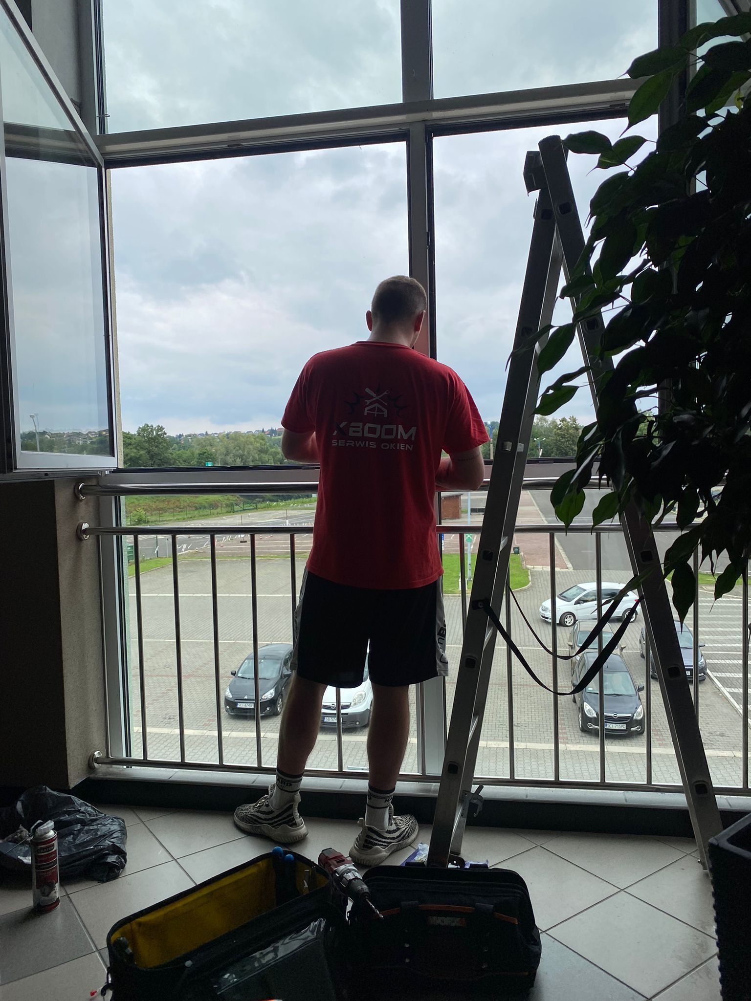Mężczyzna w czerwonej koszulce z logo firmy serwisującej okna, stojący na balkonie i regulujący otwarte skrzydło okienne, drabina oparta o ramę okienną, widok na parking i zieleń za oknem.
