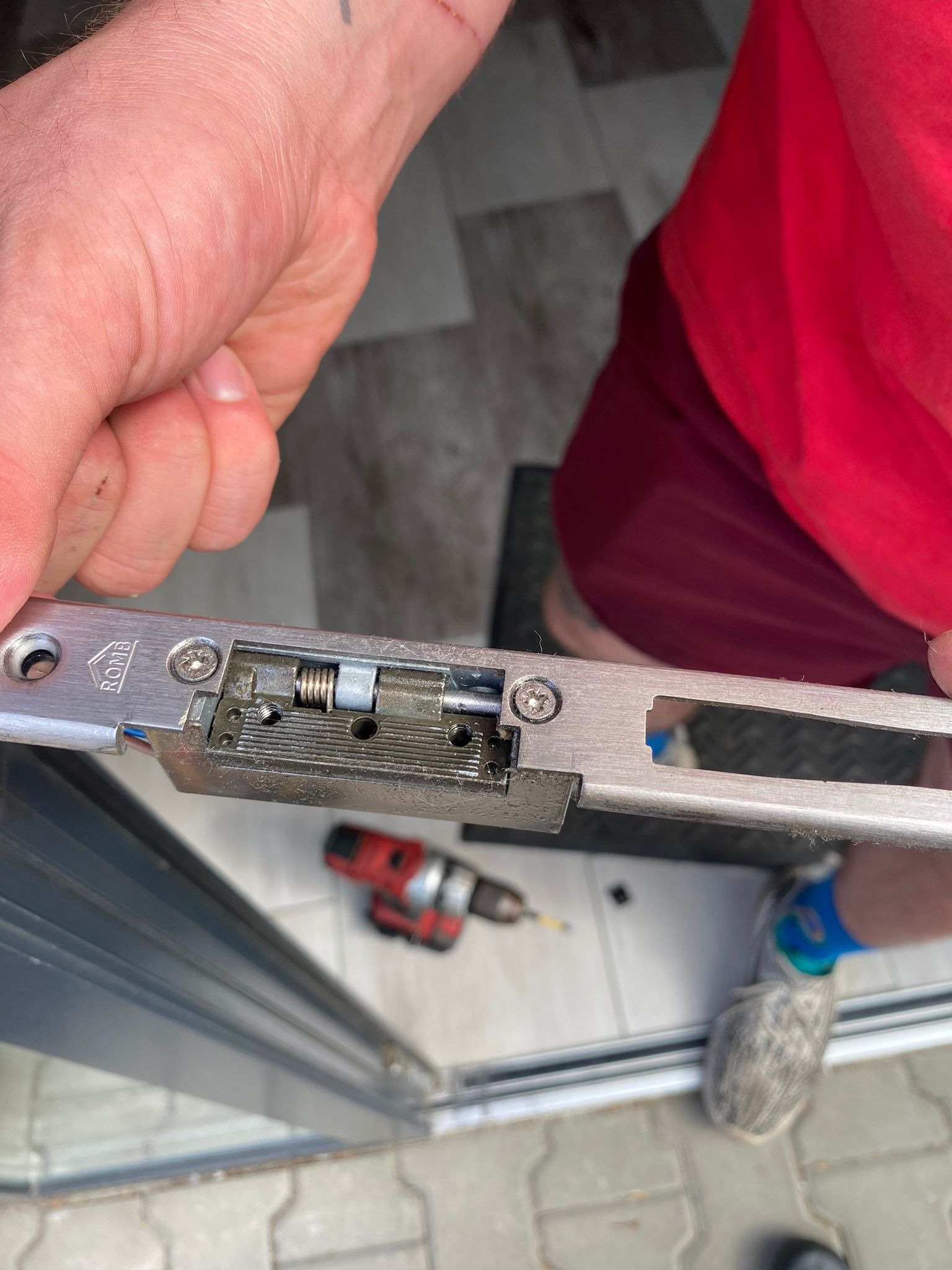 Otwarta listwa zamka drzwiowego marki ROMB, widoczny mechanizm z sprężyną, w tle wkrętarka akumulatorowa i fragment drzwi.