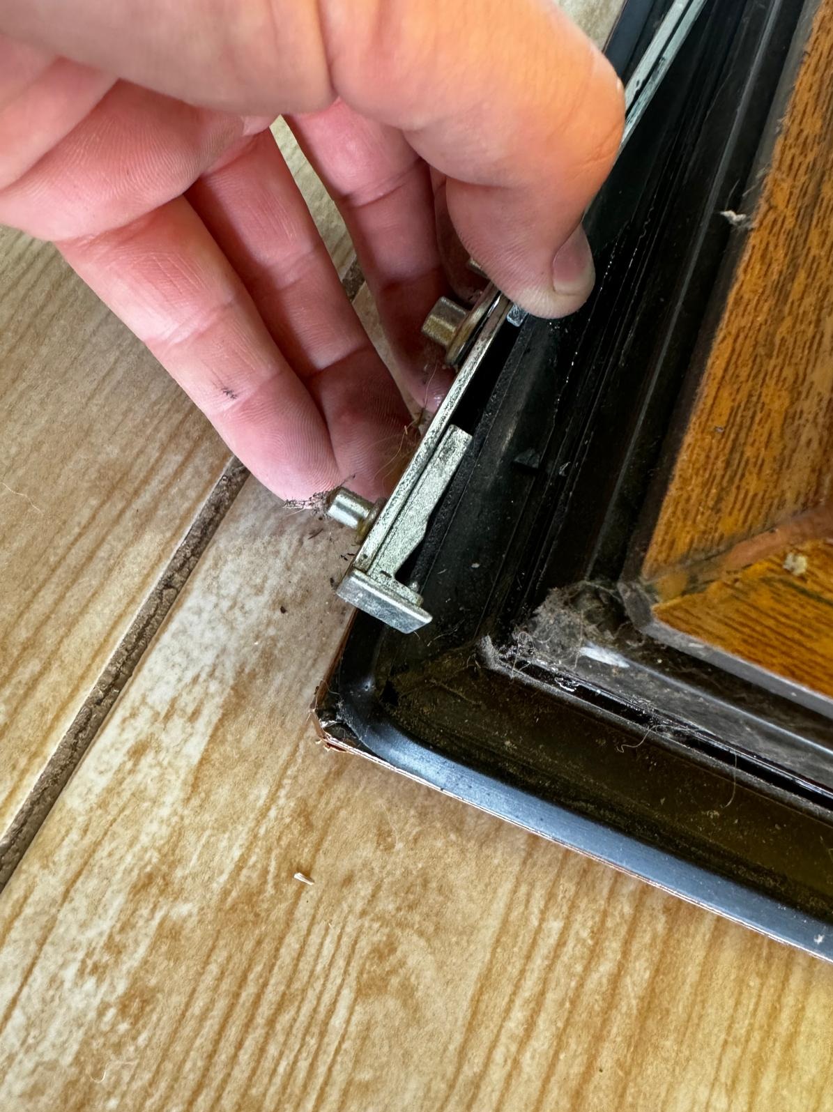 Detail ujęcia ręki przytrzymującej metalowy element regulacyjny w brudnej, drewnianej ramie okiennej w Bielsko-Białej, z widocznymi śladami kurzu i zabrudzeń.