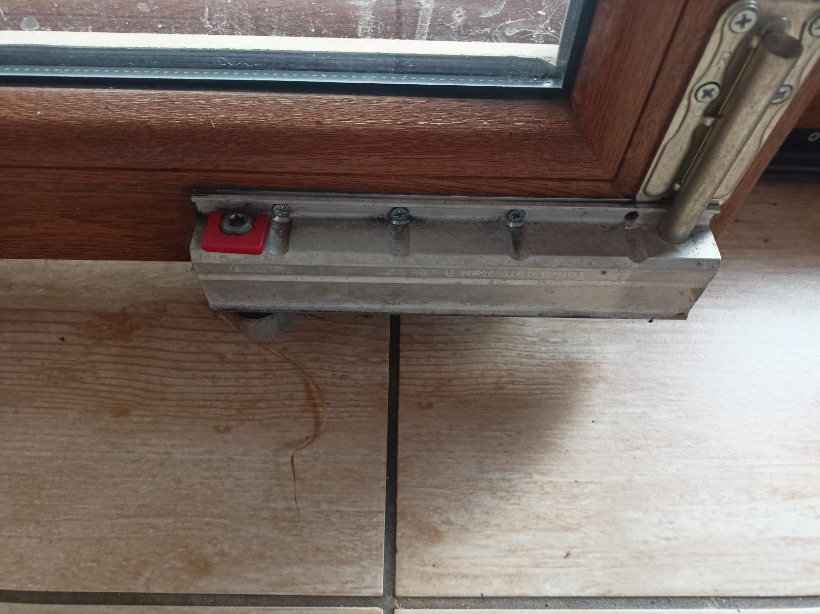 Fragment brązowej ramy okiennej z widocznym mechanizmem blokującym i uszczelką, zamontowany na kafelkach podłogowych. Widoczne ślady użytkowania i zabrudzenia na mechanizmie.