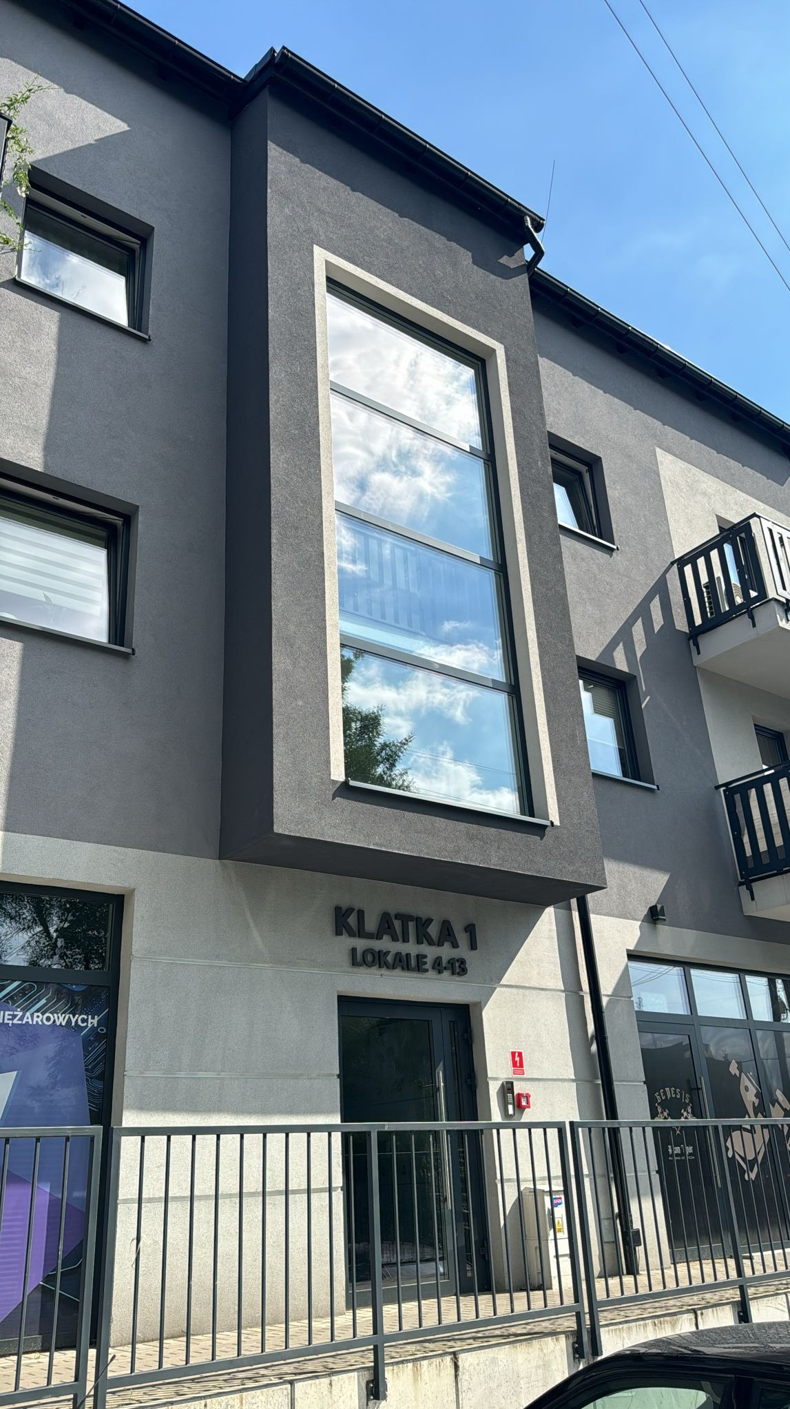 Nowoczesny budynek z dużymi oknami aluminiowymi, w których odbija się błękitne niebo z chmurami. Widoczna tabliczka z napisem 'KLATKA 1 LOKALE 4-13'.