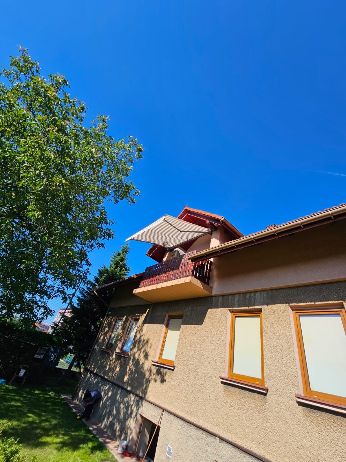 Zewnętrzny widok domu z beżową elewacją, drewnianymi oknami i balkonem z rozłożoną markizą w paski, sfotografowany w słoneczny dzień z perspektywy żabiej.