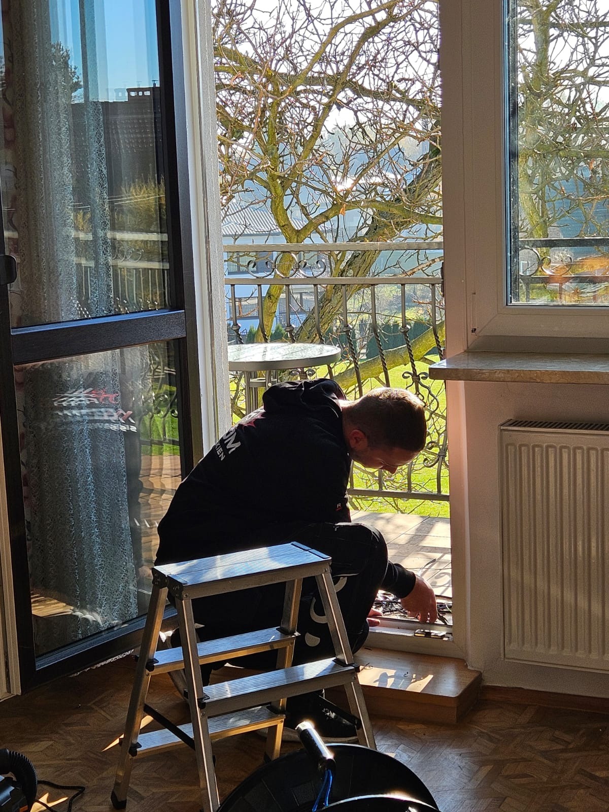 Mężczyzna w czarnej bluzie, klęczący na drabince, zajmuje się uszczelnianiem okna balkonowego z widokiem na ogród i metalową barierkę balkonu.