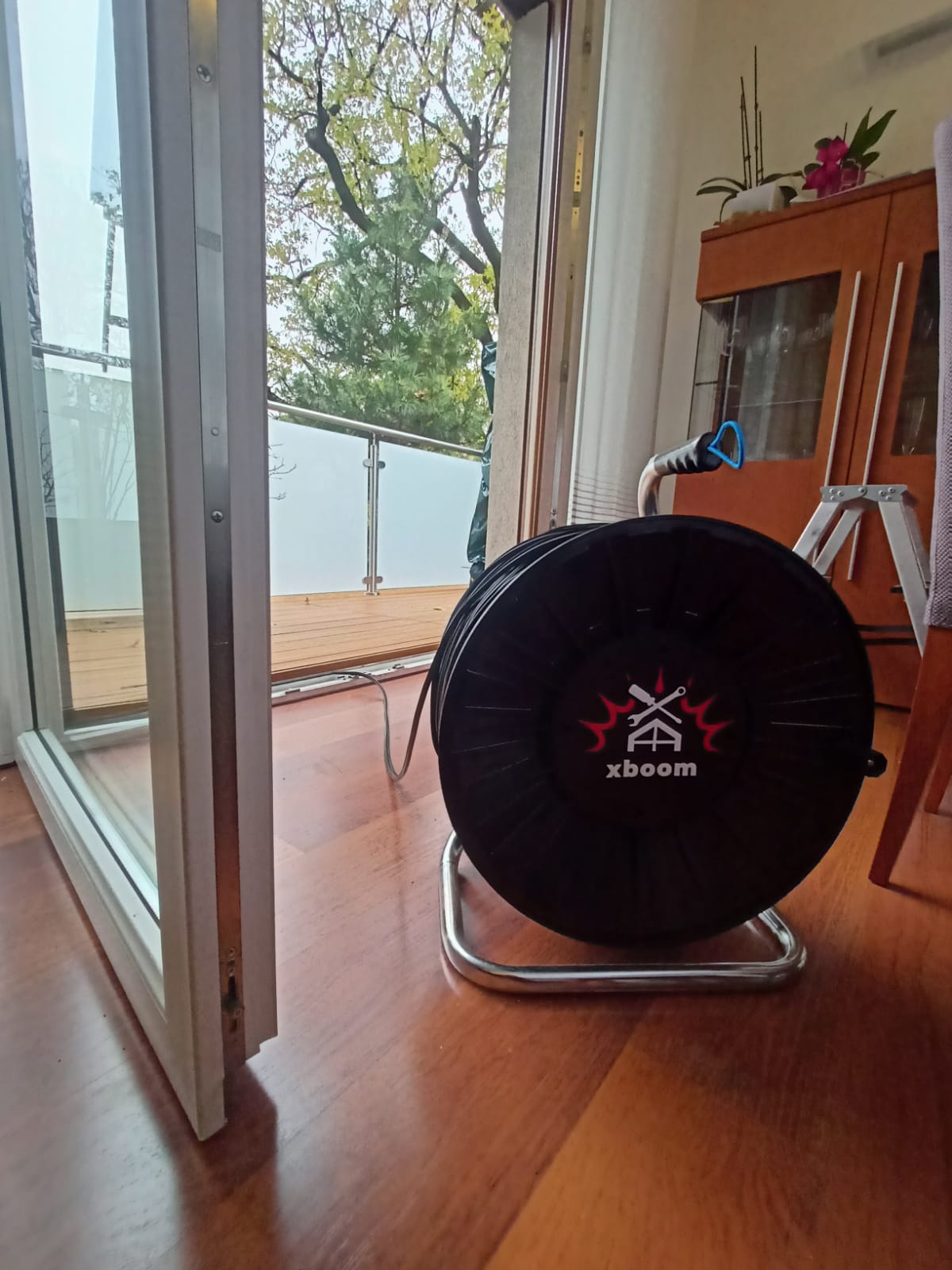 Otwarty balkon z widokiem na drzewo przez rozwarte białe okno, obok zwinięty wąż ogrodowy na metalowym stojaku z logo xboom.