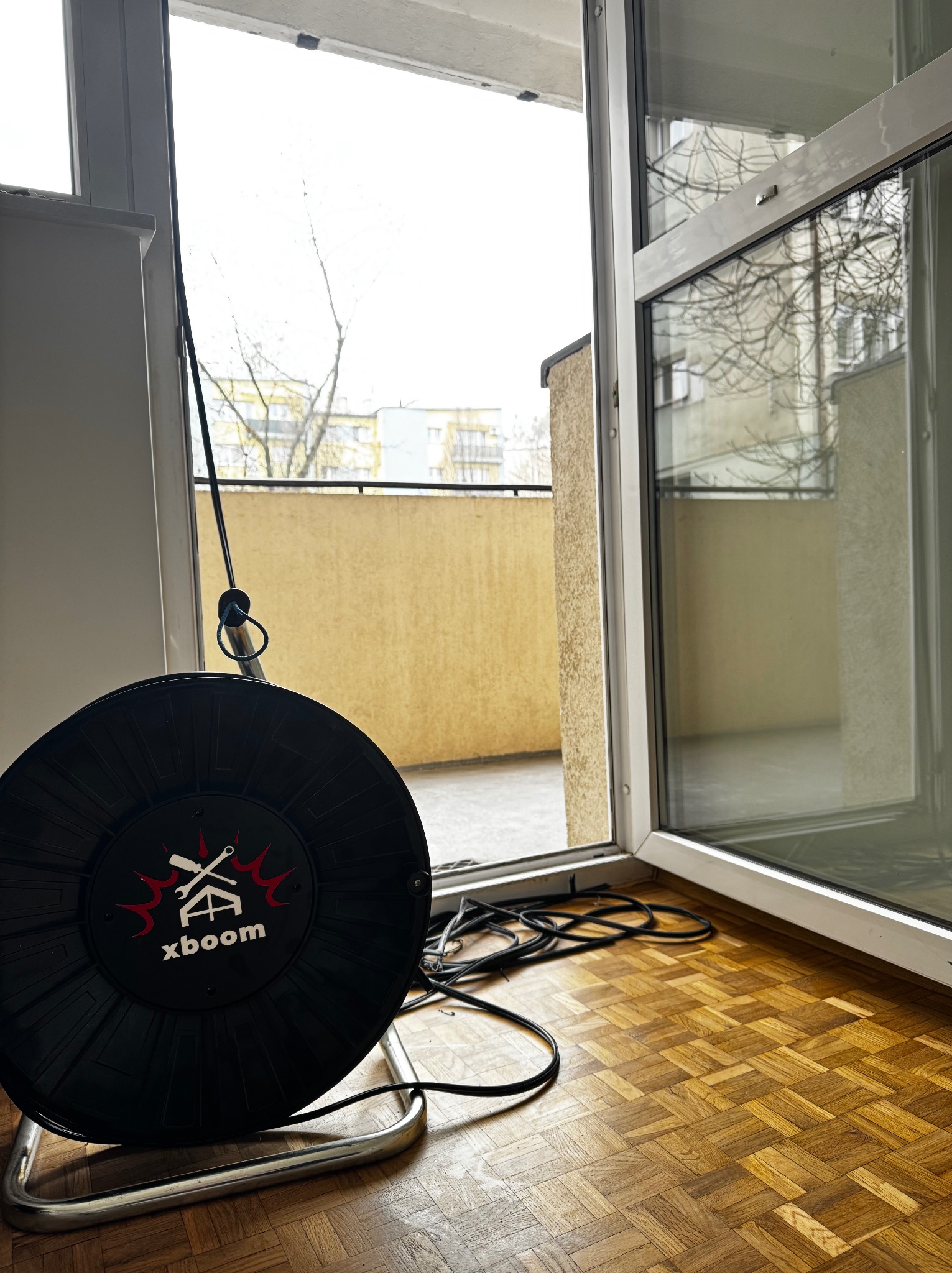 Otwarty balkon z widokiem na blok mieszkalny, widoczna zwinięta przedłużka elektryczna Xboom na podłodze z parkietu w jodełkę.