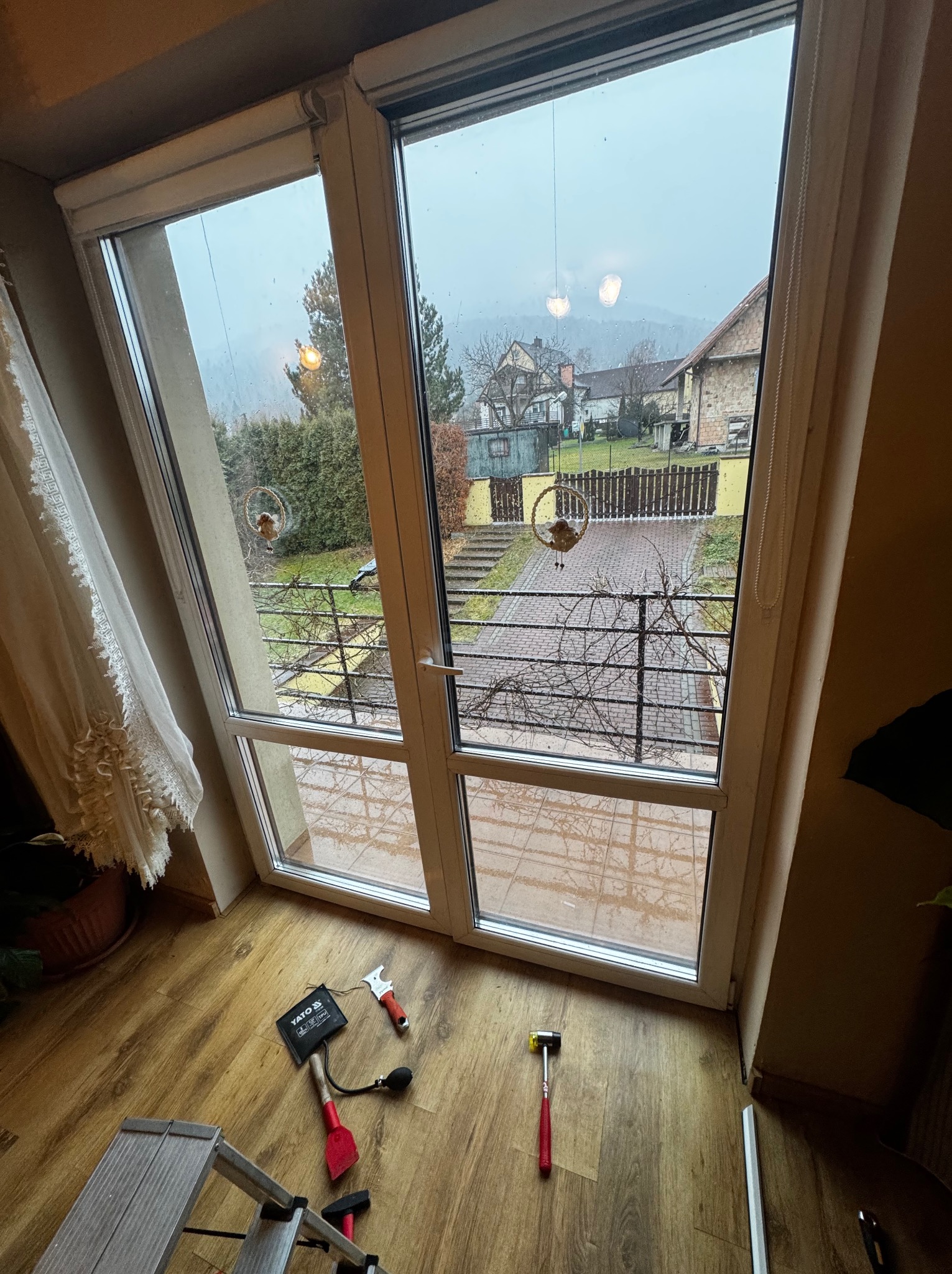 Białe drzwi balkonowe z widokiem na zewnątrz, na podłodze narzędzia: młotek gumowy, szpachelka, przyrząd do regulacji okien, drabina aluminiowa. Widoczny element dekoracyjny na oknie.