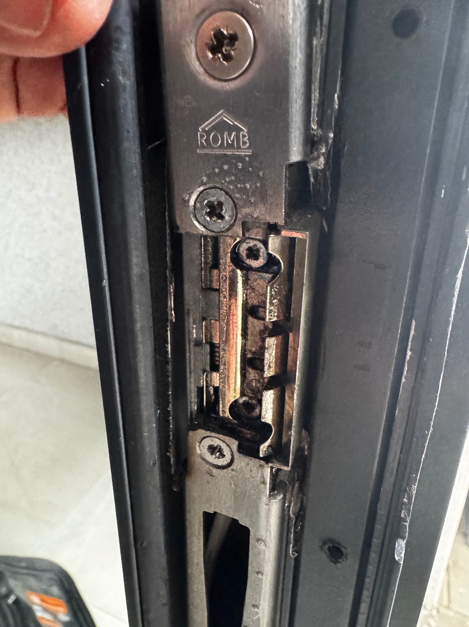 Metalowy zamek drzwiowy z widocznym mechanizmem i śrubami mocującymi, zbliżenie na element ryglowania w czarnej ramie drzwiowej, widoczne ślady użytkowania i zabrudzenia.