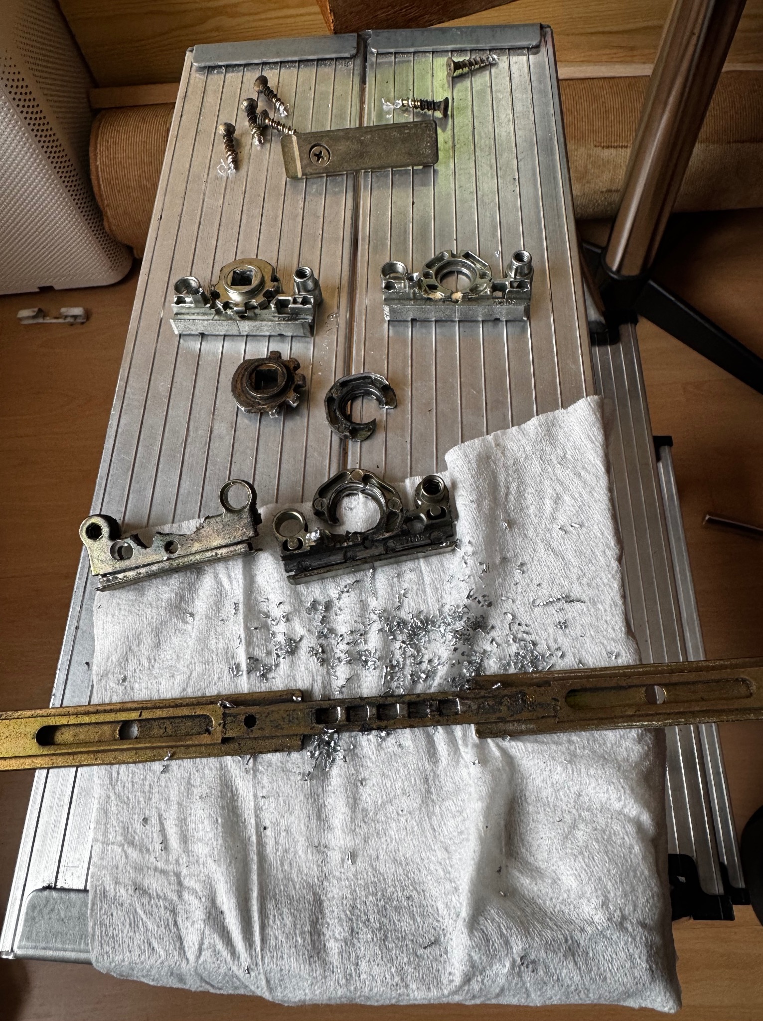 Metalowe elementy okienne, w tym pęknięta kaseta pod klamką, śruby i wióry, rozłożone na białej ściereczce na aluminiowej drabinie.