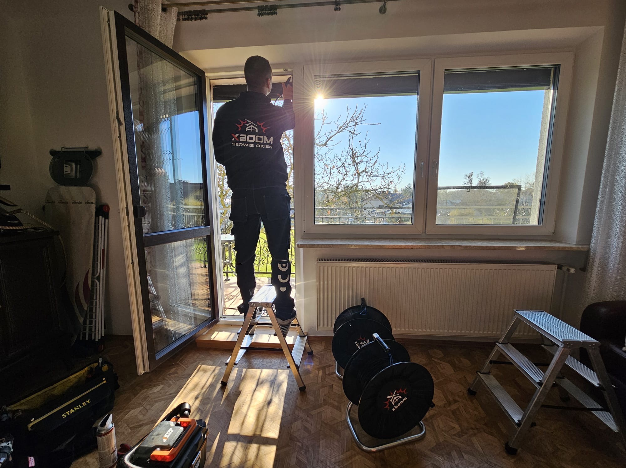 Mężczyzna w uniformie firmy XBOOM stojący na aluminiowej drabinie podczas regulacji okna PCV, widok z wnętrza mieszkania na drzewa i błękitne niebo.