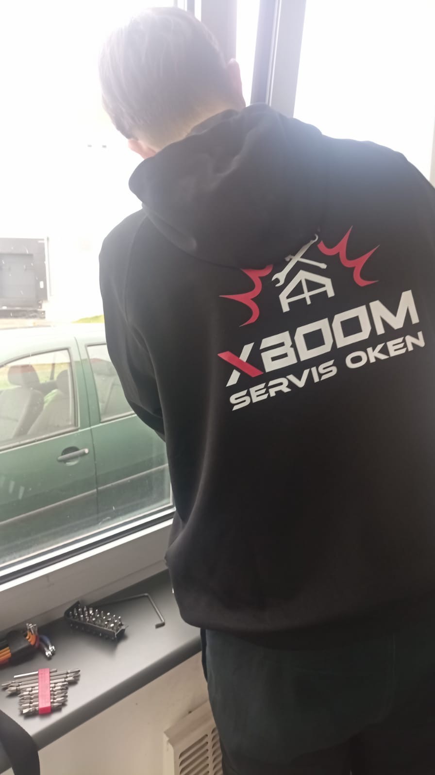 Pracownik firmy z Bielska-Białej w bluzie z logo 'XBOOM SERWIS OKIEN' stoi przy oknie, na parapecie leżą narzędzia.