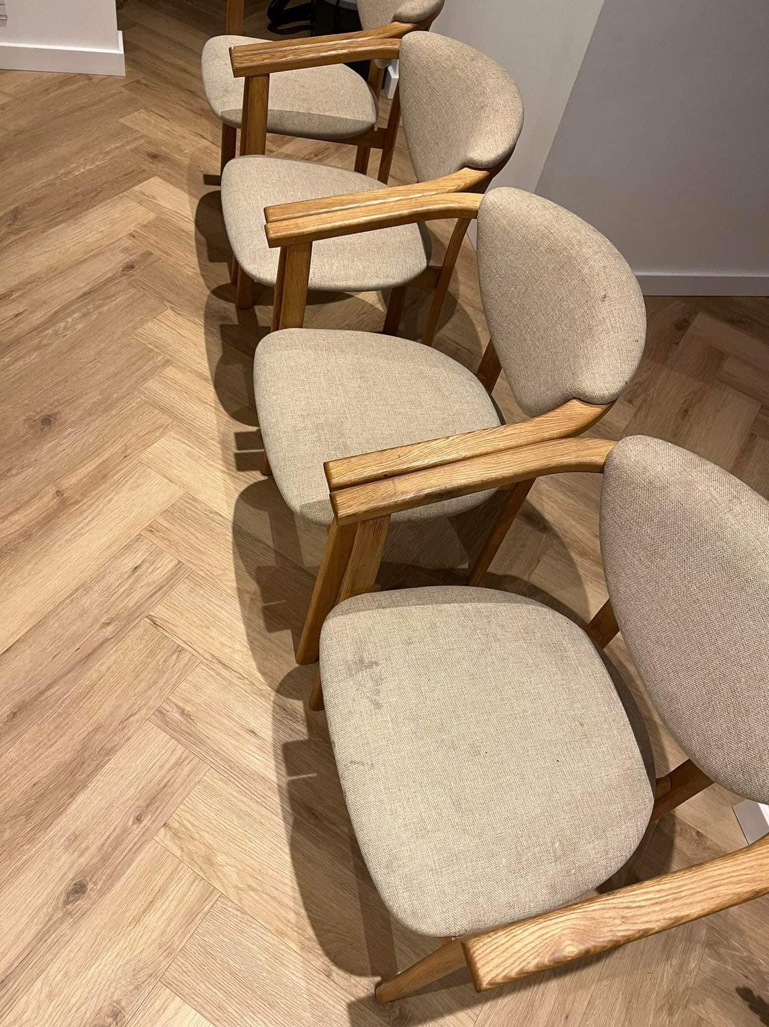 Rząd czterech krzeseł z drewnianymi ramami i beżową tapicerką na drewnianej podłodze ułożonej w jodełkę, widoczne plamy na siedzisku jednego z krzeseł.