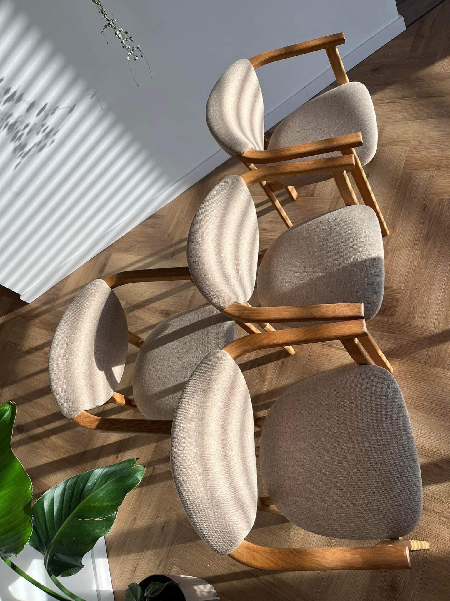 Pięć krzeseł z jasną tapicerką i drewnianymi ramami ustawionych na parkiecie w jodełkę, oświetlonych promieniami słonecznymi przez żaluzje, z widoczną rośliną doniczkową w lewym dolnym rogu.