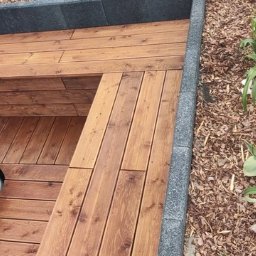 Holz - Brązowy taras z drewna z wbudowanym siedziskiem i obrzeżem z szarej kostki. Obok rabata z kwiatami, w tle widoczny fragment jacuzzi.