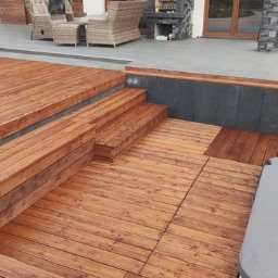 Holz - Brązowy taras z drewna z kilkoma poziomami schodów, obok szarej wanny z hydromasażem i szarej podłogi z meblami ogrodowymi w tle.