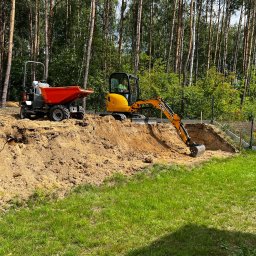 VNUK - Żółta mini koparka JCB i wywrotka na wykopanym terenie. W tle wysokie drzewa, częściowo ogrodzenie. Prace ziemne w toku, widoczny wykop.