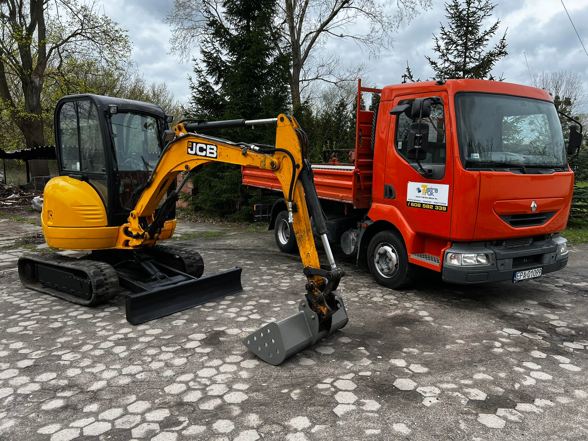 Żółta mini koparka JCB z łyżką i lemieszem stoi na kostce brukowej obok pomarańczowej wywrotki z widocznym logo firmy na drzwiach.