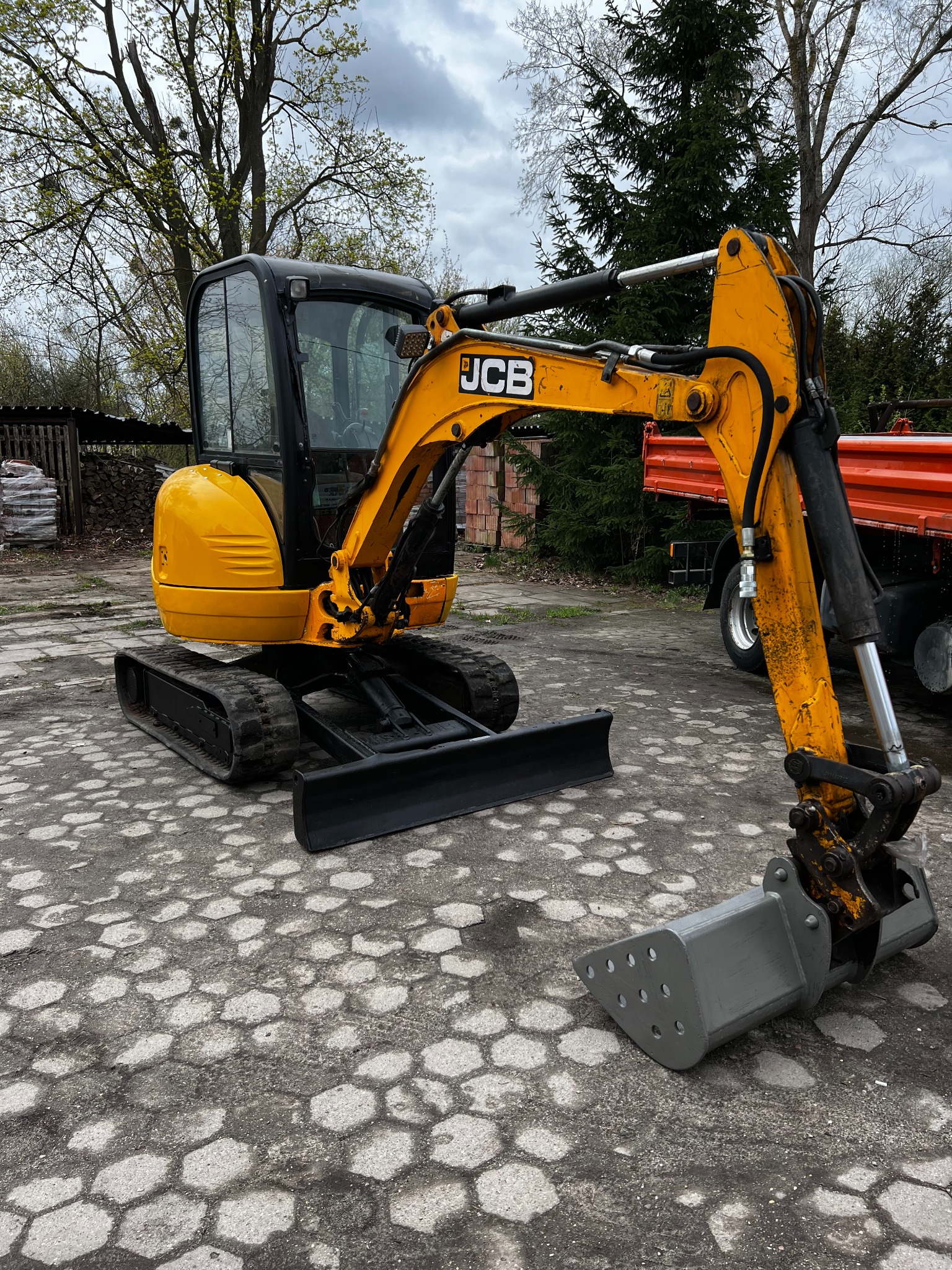 Żółta minikoparka JCB na placu z kostki, z pomarańczową wywrotką w tle. Widoczne ślady użytkowania na maszynie. Pochmurne niebo w tle.